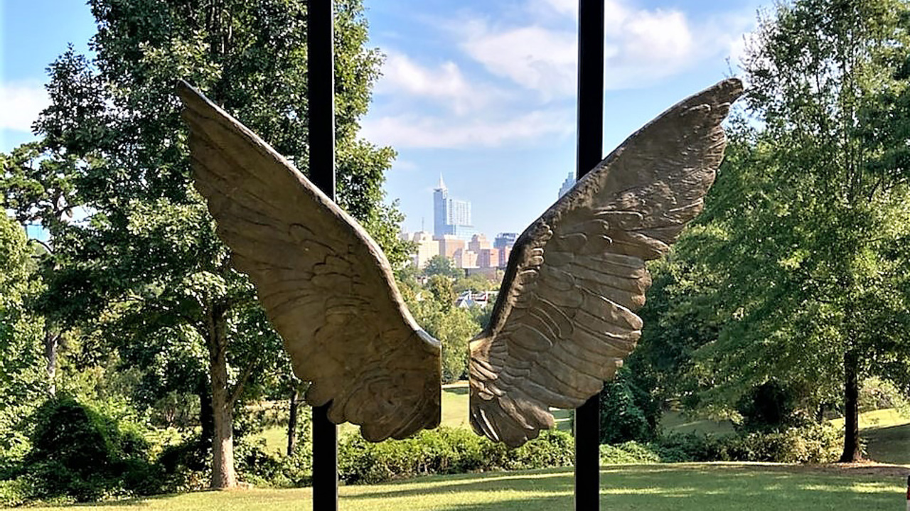 a large pair of gray wings suspended on black poles with a cityscape framed in the background between the wings, surrounded by greenery and trees. the sculpture is by Jorge Marín