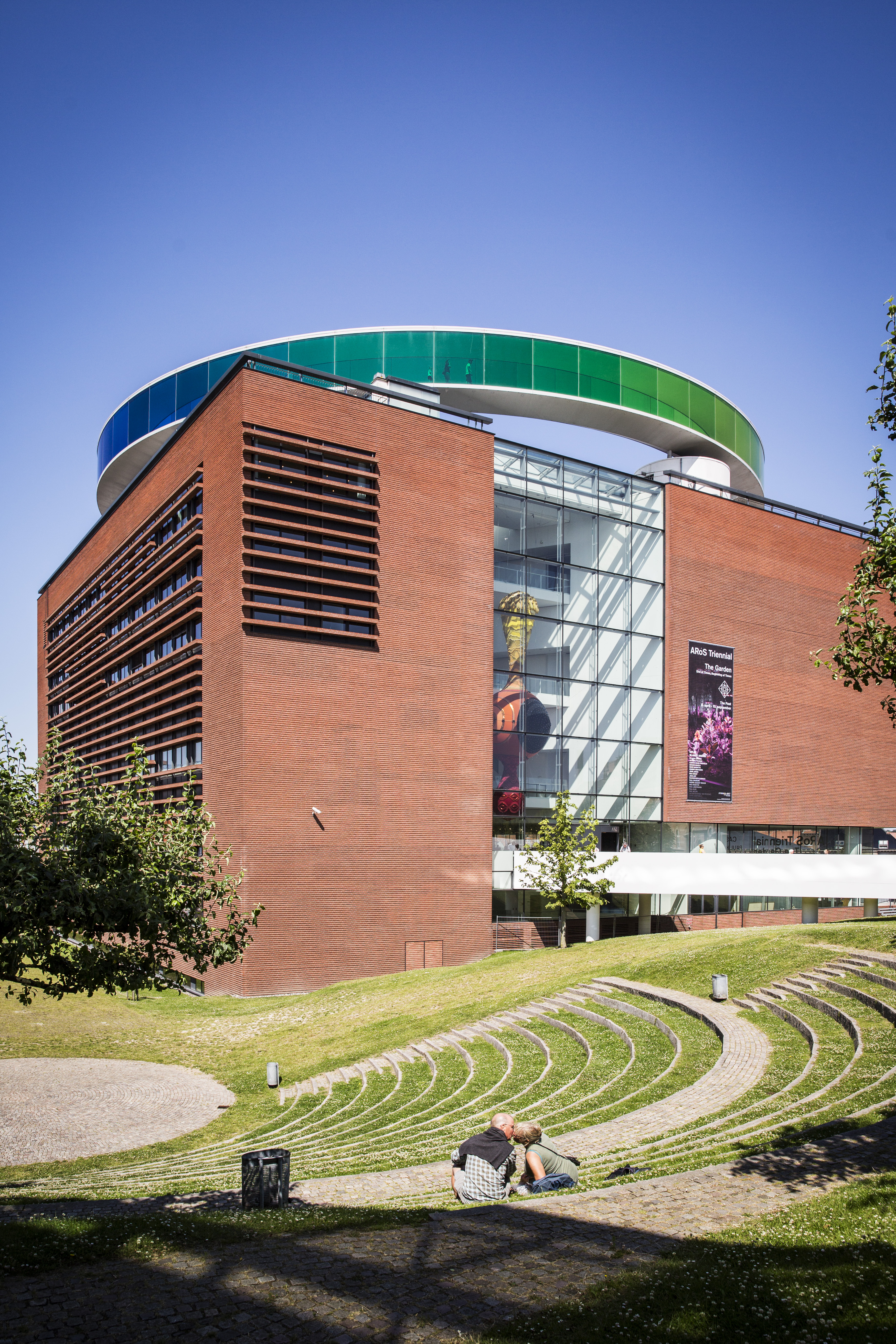 Square, red-bricked museum building with a circular, rainbow walkway on the roof.