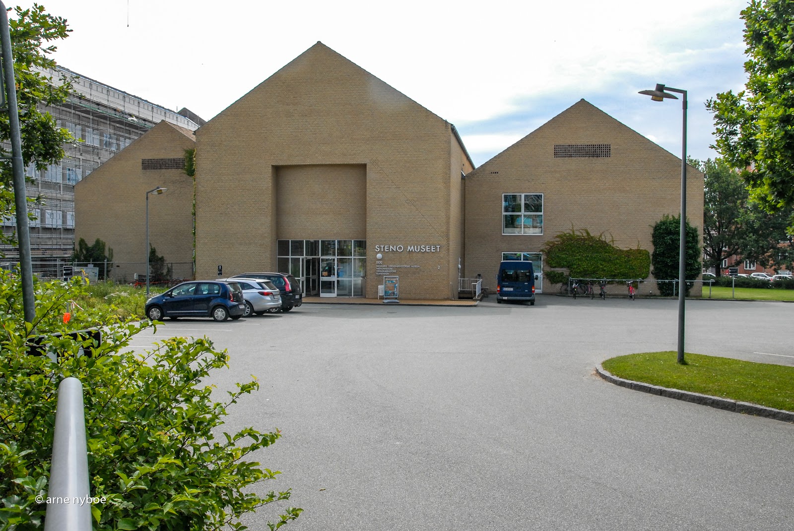 Modern Steno Museet building with tan brick facades and triangular rooflines, featuring a paved parking area with cars in front.
