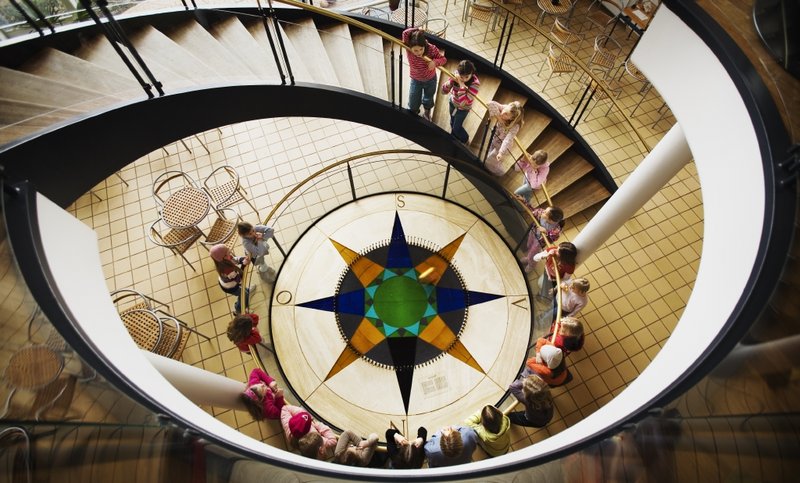 Aerial view of a spiral staircase with black railings curving around a decorative compass rose mosaic floor, with people and wicker chairs visible below.